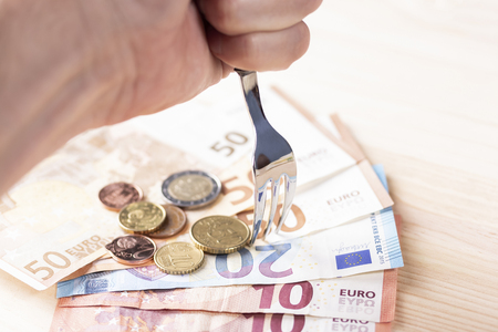 A shiny metal fork pierces a pile of euro banknotes and coins on a wooden table. The concept of...