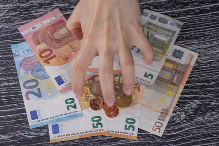 Twisted by greed, the hand of the woman rakes up a bunch of Euro notes and coins to yourself at a dark wooden table. The concept of selfishness and fraudの写真素材