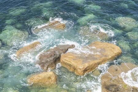 Ocean coast. Rocks and stones are washed by turquoise foamy waves of the bay on a sunny day with a clear blue skyの写真素材