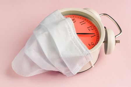 Alarm clock in a medical disposable mask on a pink background with a red dial. The concept of...
