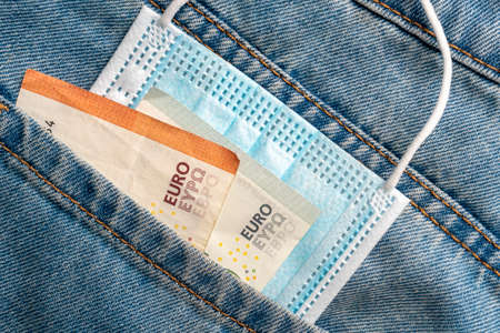 Close-up of a protective medical mask and money in the pocket of old jeans on a wooden background....