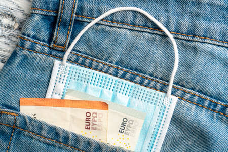 Close-up of a protective medical mask and euro banknotes in the pocket of blue old jeans on a gray...