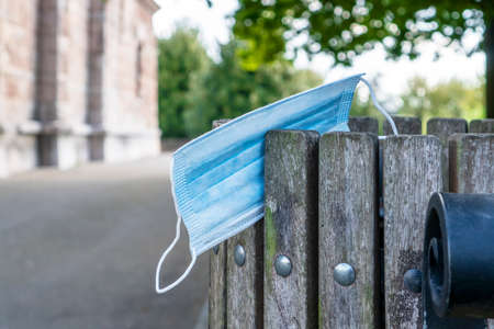 Thrown used medical protective mask on the edge of a wooden trash can. Building and foliage of trees in blur. Close-up. Ecology pollution conceptの写真素材