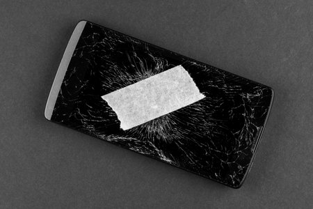 Broken mobile phone with shattered screen glass and masking piece of sticky paper tapeの写真素材