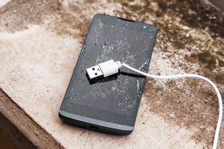 Shattered glass touchscreen of a black broken smartphone with a white charging cable on exterior...
