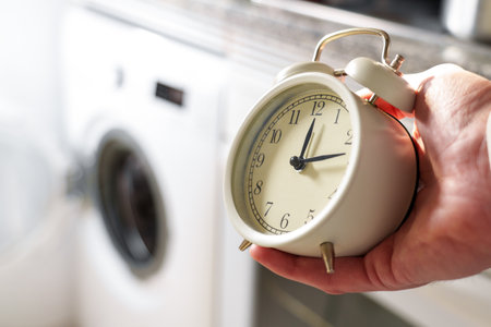 Alarm clock in hand and washing machine on the background close up. Energy saving and warranty period conceptの写真素材