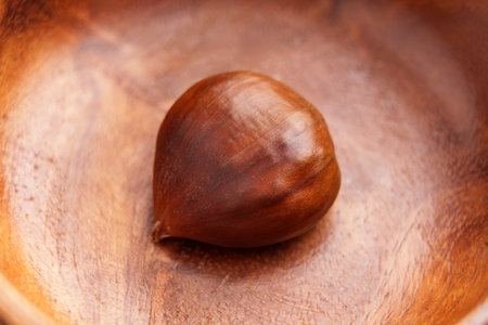 A single chestnut in smooth wooden bowl. Brown nut surface on textured grainy wood. Close up