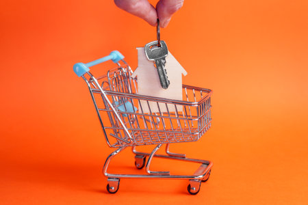 Hand places bunch of keys with wooden house keychain into mini shopping cart. Bright orange background. Side view. Real estate and home ownership conceptの写真素材