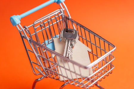 Home keys in small shopping cart. Bright creative orange color background. Keychain with wooden house shape. Conceptual miniatureの写真素材