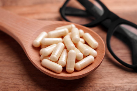 Beige capsules in wooden spoon on brown textured background with black glasses. Medicine study...