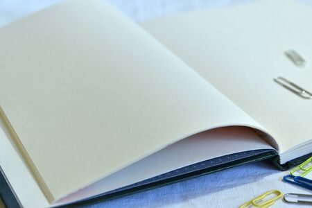 A green sketchbook and multi-colored paper clips on a white fabric tablecloth in cool colors with...