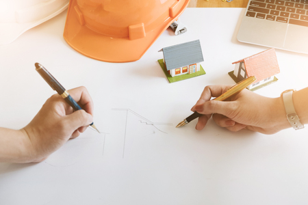 Architect team freehand drawing with pen on blueprint architectural in workplace, laptop small house modell and safety helmet on table, Construction Concept.の写真素材