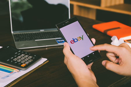 CHIANG MAI,THAILAND - July 7,2018: Man holds Mobile Phone using Ebay app on the screen with computer laptop on wooden table.のeditorial素材