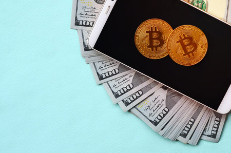 Bitcoins lies on a smartphone and hundred dollar bills on a light blue background.の写真素材