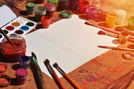 Background image showing interest in watercolor painting and art. A blank sheet of paper, surrounded by brushes, cans with watercolor paint and gouache, which lie on an old and stained wooden tableの写真素材