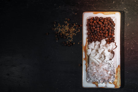 Sea salt flakes and pepper in dish has peppercorns pile and oregano on black wood background, top view, copy space, condiment food concept.の写真素材
