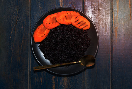 Black rice in plate with vegetable carrot has spoon isolated on blue wood background close up, top view, healthy food and drink concept.の写真素材