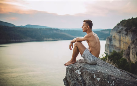Young man sitting on the edge of the cliff near the lake after swimming and looking far awayの素材
