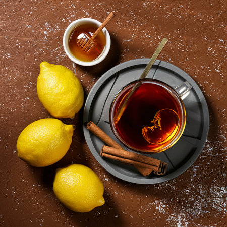 Black tea in glass cup served with fresh lemons, honey and cinnamon sticks over a brown texture backgroundの素材