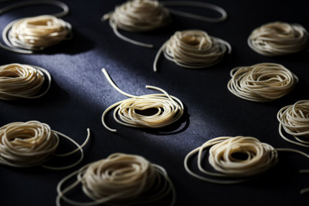 Several small, coiled nests of uncooked spaghetti are artfully arranged on a dark, textured surface. Dramatic light and shadow create a visually appealing composition.の素材