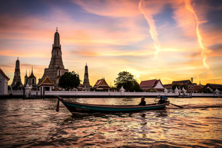 A traditional longtail boat with a lone passenger glides across the Chao Phraya River. The iconic Wat Arun temple complex is beautifully illuminated by the warm hues of a dramatic sunset.の素材