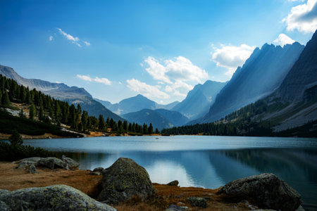 A tranquil alpine lake mirrors the surrounding rugged mountain peaks and a scattering of white clouds. Lush green pine forests line the shore, with rocky terrain in the foreground.の素材