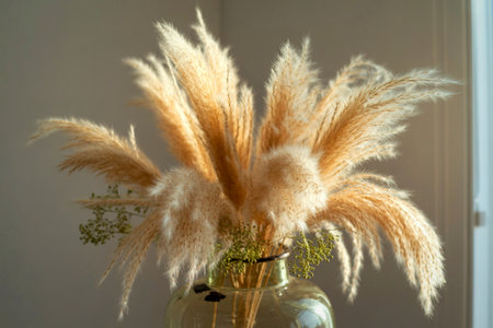 A bouquet of dried pampas grass, in varying shades of beige and off-white, sits in a clear glass...