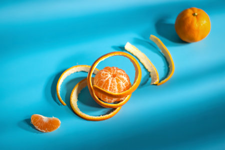 A whole clementine sits above a peeled clementine with its skin artfully arranged around it, on a vibrant blue surface with shadows.の写真素材