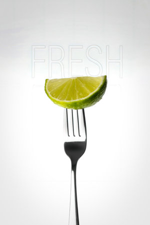 A vibrant green lime wedge is precisely balanced on the tines of a metal fork, presented against a clean white backdrop.の写真素材