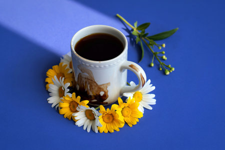 A white mug of dark coffee sits on a blue surface, encircled by a vibrant wreath of daisies and yellow wildflowers, with a sprig of greenery.の写真素材