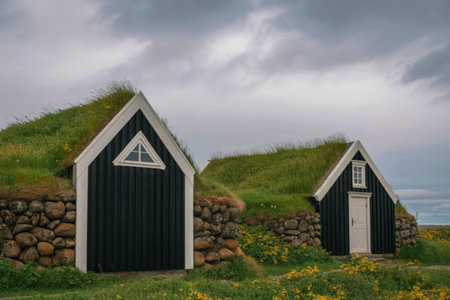 Two historic Icelandic turf houses with dark facades and grass-covered roofs stand on a hillside overlooking the sea.の写真素材