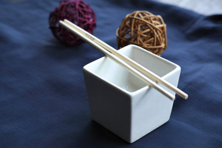 A pair of wooden chopsticks lies across a white ceramic square holder. Two decorative balls are blurred in the background.の写真素材