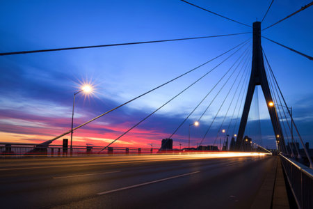 A striking view of a modern cable-stayed bridge at dusk, featuring a vibrant sunset and blurred car lights creating a sense of motion.の写真素材