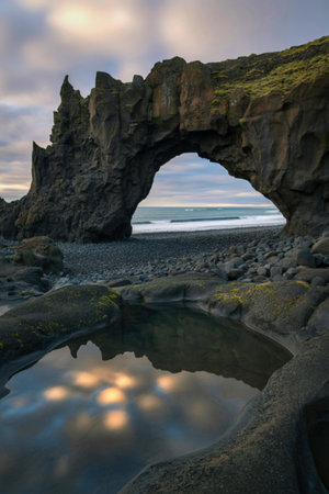 A natural stone archway frames a distant ocean view, with a calm pool reflecting the sky and sunlight on the rocky shore.の写真素材