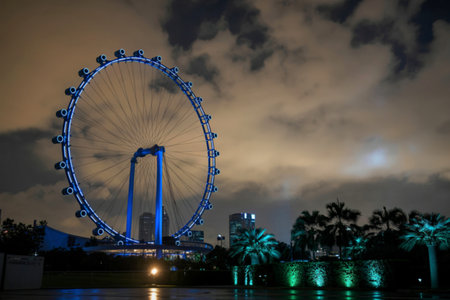 The Singapore Flyer observation wheel glows with blue lights against a dramatic cloudy night sky, reflecting on the water below.の写真素材