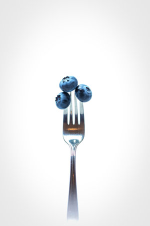 A close-up shot shows three ripe blueberries skewered on the tines of a shiny silver fork, isolated on a clean white backdrop.の写真素材