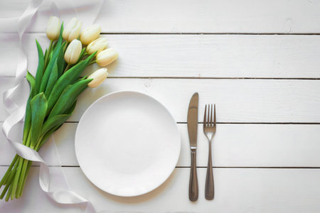 A white plate, silverware, and a bouquet of white tulips are arranged on a white wooden table, suggesting a special occasion.の写真素材