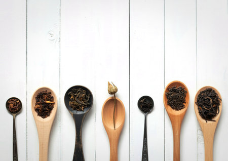 A collection of wooden and metal spoons, each holding a different variety of loose leaf tea, arranged on a white wooden surface.の写真素材
