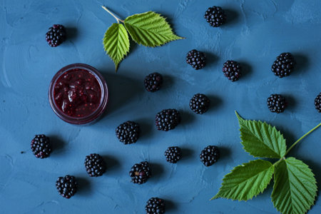 A close-up image showcasing fresh blackberries scattered around a jar of blackberry jam, accompanied...