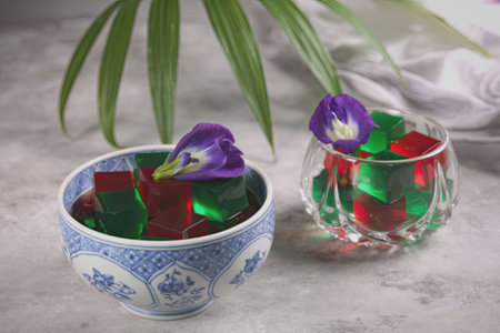 Two decorative bowls, one blue and white and the other clear, filled with colorful glass stones and purple flowers, placed on a light-colored surface with a green plant in the background.の写真素材