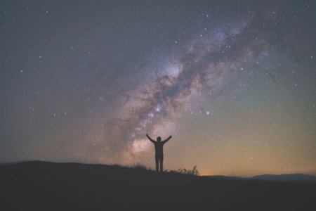 A person stands with arms raised, silhouetted against the vibrant Milky Way galaxy, with distant...
