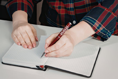 A person wearing a red and blue plaid shirt is writing in a notebook with a red pen.