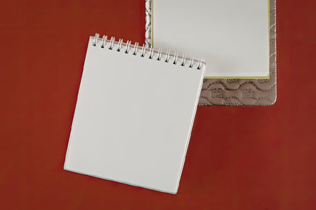 A spiral bound notebook with blank pages is placed on a red surface, with another notebook partially...