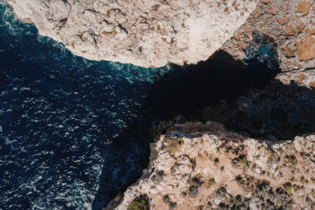 An aerial view of a rugged rocky coastline with a deep blue ocean and clear shadow on the land.