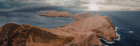 An aerial view of a red rocky island surrounded by blue water, with the sun shining through clouds...