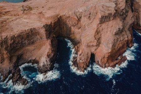 This aerial image captures the rugged beauty of red rock cliffs towering over the deep blue ocean....