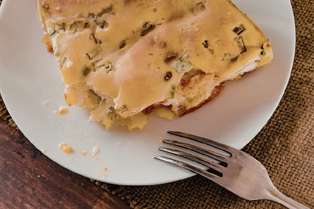A slice of pizza on a white plate with a fork, placed on a wooden table with a brown placemat.