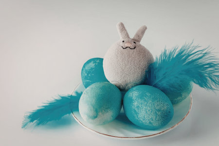 An adorable white bunny surrounded by blue eggs on a white plate with feathers, symbolizing Easter...
