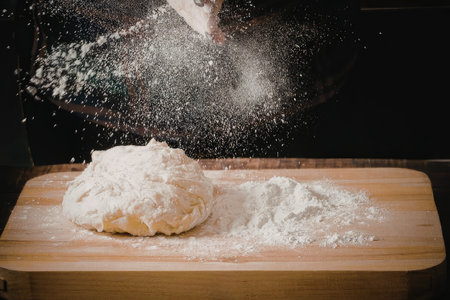 A ball of dough and a pile of flour on a wooden cutting board with flour being sprinkled over it.