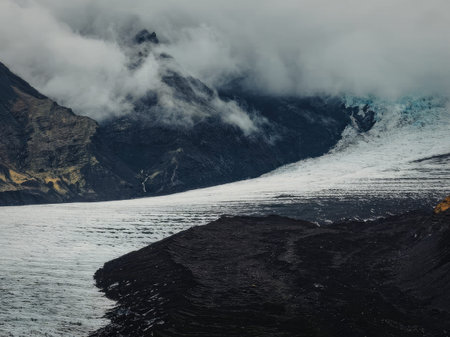 A glacier extends from the mountains onto dark land under a cloudy sky